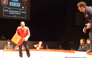Capture d'écran de la vidéo suggérée dans l'article : on y voit 2 joueurs, un rouge et un bleu, sur la scène, ainsi que le panneau de scores et le titre de l'impro.